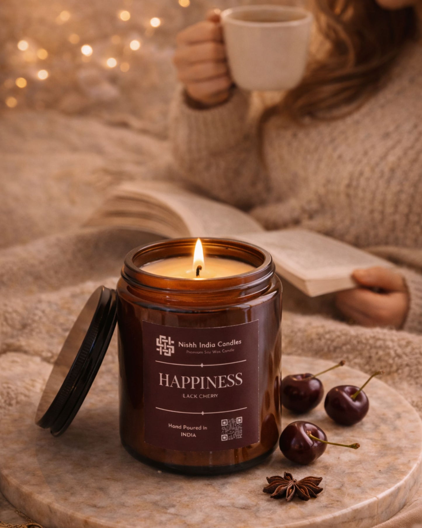 Nishh India Happiness Black Cherry scented soy candle lit in cozy home setting with woman reading and sipping coffee in background, warm ambient luxury decor.