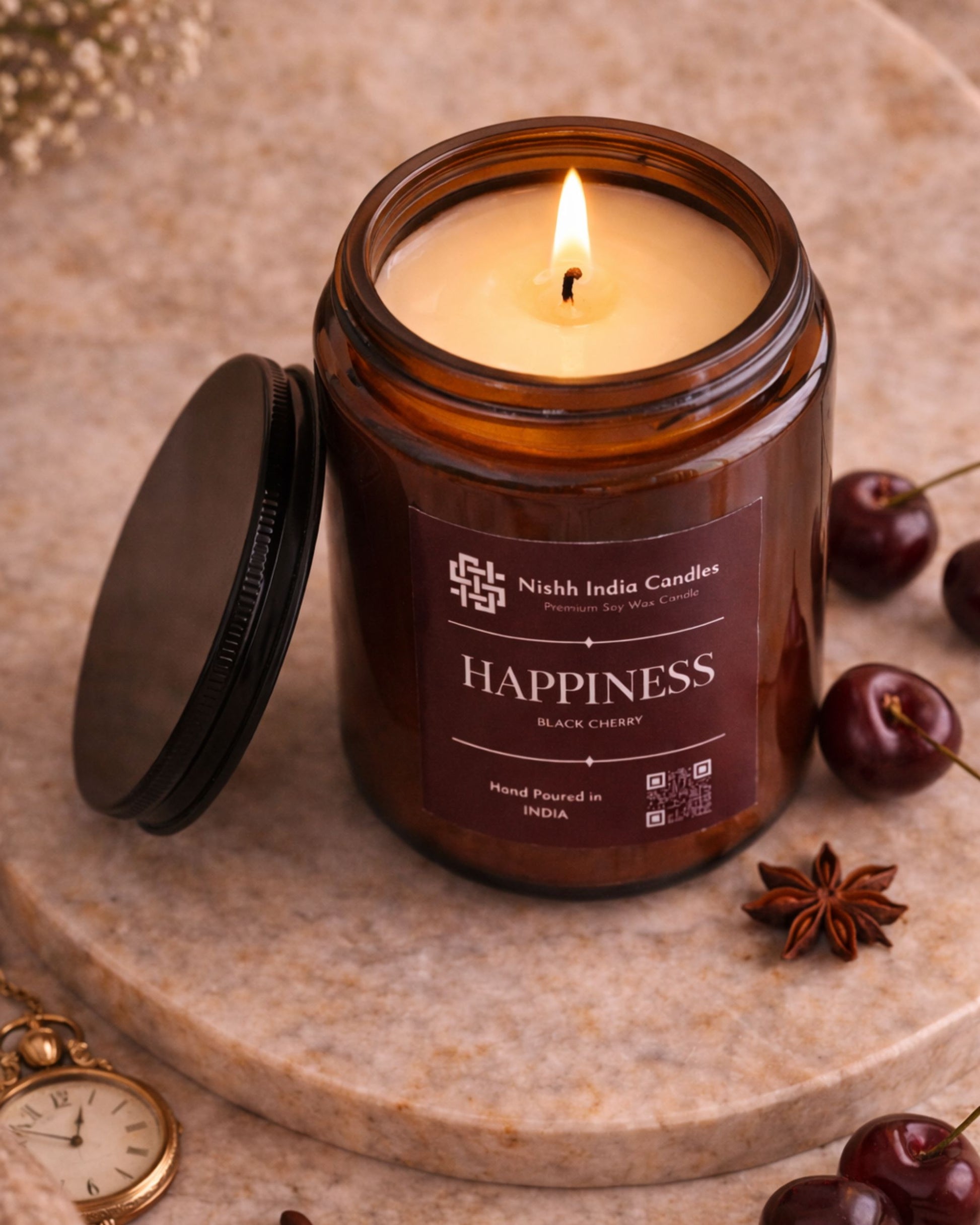 Lit Nishh India Happiness Black Cherry soy wax candle in amber glass jar styled on marble surface with cherries and warm luxury decor ambience.