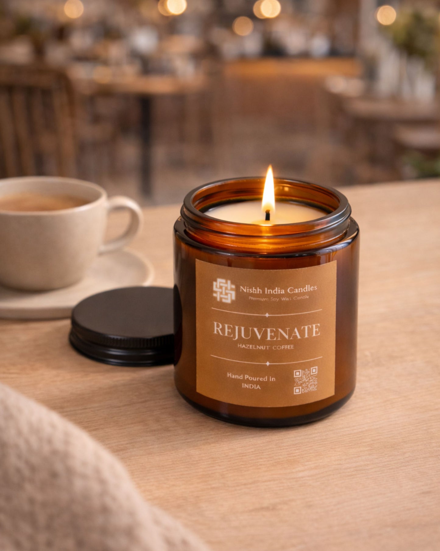 Nishh India Rejuvenate Hazelnut Coffee scented candle lit on cafe table with blurred warm café background creating cozy atmosphere